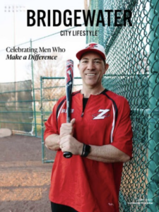 An image of Coach Duke Baxter holding a bat in front of a baseball field's chain link fence. He's smiling, wearing a white cap and red t-shirt emblazoned with a Z for his company, Zoned Sports