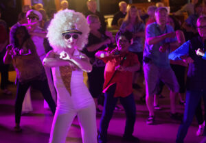 A dance with a woman in a white bodysuit and large blonde afro in the foreground. Lights add a violet hue to the scene